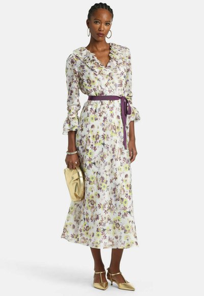 Femeie purtând o rochie midi cu mâneci lungi cu imprimeu floral, guler și mâneci cu volane, centură mov, pantofi cu toc auriu și ținând o plic auriu.