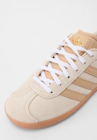 Beige mockasin sneaker med vita snören, beige detaljer och tre ränder. Har en strukturerad gummisula och en gul logotyp på tungan.