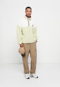 Light green fleece pullover with a white chest section, half-zip, maroon accents. Paired with beige cargo pants and a beige cap.