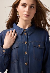 Camicia di denim con colletto, bottoni frontali e due tasche sul petto. Presenta cuciture chiare e una texture liscia.