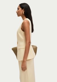 Chaleco entallado color crema con diseño de peplum, llevado sobre pantalones de pierna ancha a juego. La modelo sostiene un bolso de mano marrón. Textura suave.