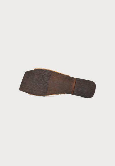 Semelle d'une sandale plate avec une texture boisée et un talon et un bout légèrement carrés, présentée sur un fond clair uni.