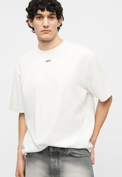 Νέος άνδρας με σγουρά σκούρα μαλλιά, φορώντας λευκό φαρδύ T-shirt με το κείμενο "OFF" και ξεθωριασμένο γκρι τζιν, στέκεται μπροστά σε απλό φόντο.