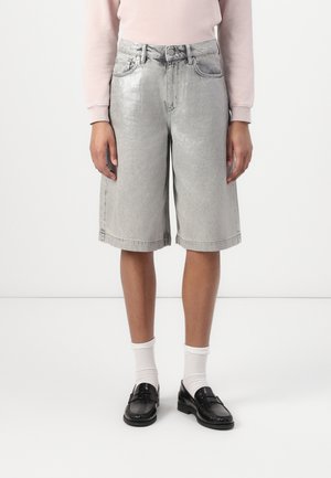 Person iført sølvmetalliske denimshorts til knæene, lyserød langærmet top, hvide sokker og sorte loafers på hvid baggrund.