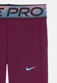Leggings Nike Pro en bordeaux, avec une ceinture multicolore ornée de lettres noires audacieuses et un petit logo Nike bleu sur la jambe.