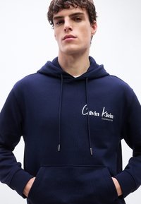 Νέος άνδρας που φοράει μια μπλε σκούρα κουκούλα Calvin Klein με λογότυπο στο στήθος, τα χέρια στην μπροστινή τσέπη και κορδόνια να κρέμονται.