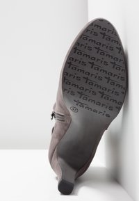Stivale alla caviglia grigio in suede con zip laterale. La suola è in gomma nera con il marchio "Tamaris" in un motivo ripetuto. Taglia 37.