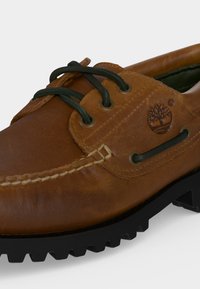 Chaussure bateau en cuir marron avec lacets noirs, coutures blanches, semelle noire robuste et logo arbre Timberland embossé sur le côté.