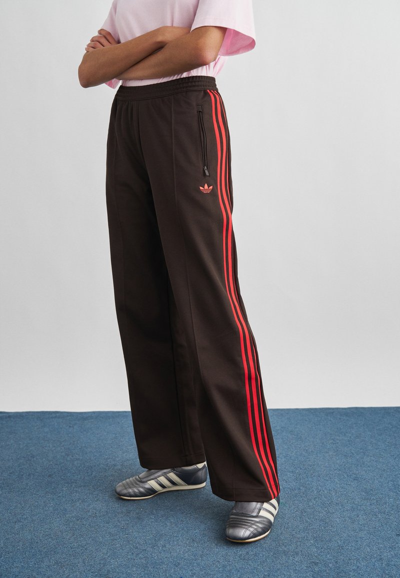 Άτομο που φοράει μαύρη φόρμα Adidas με κόκκινες ρίγες και γκρι παπούτσια Adidas, στέκεται σε μπλε χαλί με σταυρωμένα τα χέρια.