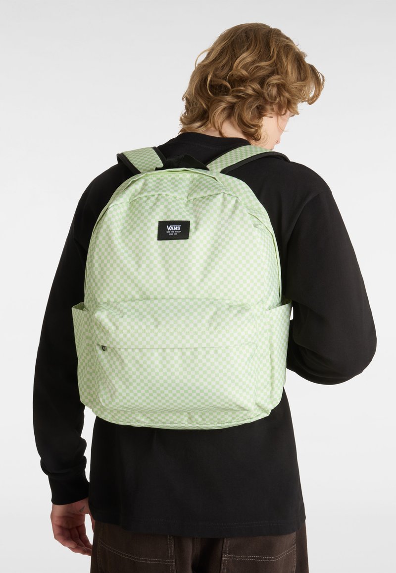 Sac à dos à carreaux verts en tissu, avec une poche avant et des bretelles rembourrées. Patch du logo de la marque en haut. La texture semble lisse.