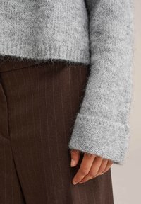 Svijetlosivi pleteni pulover s labavim krojem i rebrastim rukavima, uparen s tamnosmešnim hlačama s prugicama. Vidljivi su detalji teksturirane tkanine.