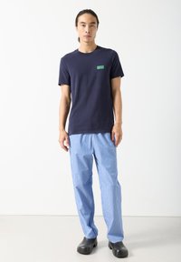 Muž s dlouhými vlasy v tmavě modrém tričku "American Vintage", modrobíle pruhovaných kalhotách a černých nazouvacích botách stojící před jednobarevným bílým pozadím.