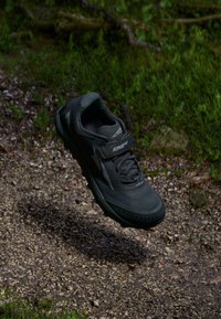 Black athletic shoe with textured upper, laces, and velcro strap, featuring a rugged sole for traction, hovering above a gravel path.