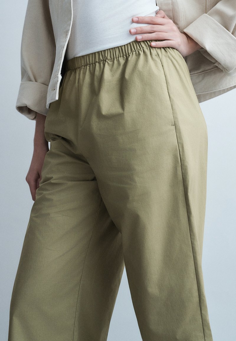 Άτομο που φοράει μπεζ παντελόνι με ελαστική μέση, λευκό ριγωτό τοπ και ανοιχτό μπεζ μπουφάν με μανίκια που έχουν γυριστεί, με το χέρι στη μέση.