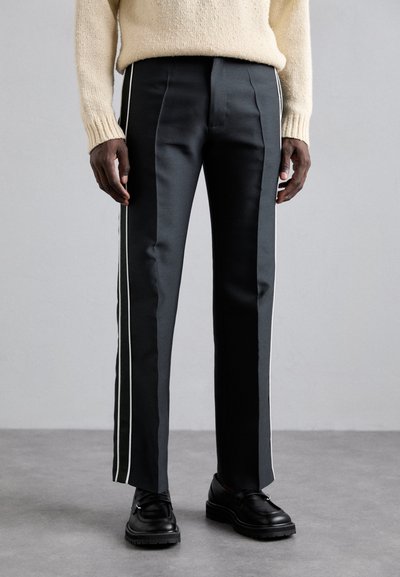 Maßgeschneiderte schwarze Hose aus einem glatten Stoff mit weißen Seitenstreifen, gerade geschnitten und mit einem sauberen Finish. Getragen mit schwarzen Schuhen.
