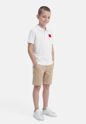 Zēns stāv ar rokām kabatās, valkājot baltu polo krekliņu ar sarkanu logo, bēšas šortus un baltas sporta kurpes uz baltas fona.