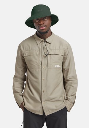 Uomo con cappello verde scuro a tesa larga e camicia beige con bottoni e tasche sul petto con logo, mani nelle tasche, rivolto in avanti.