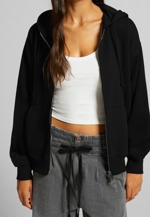 Femme portant un sweat à capuche noir zippé sur un débardeur court blanc avec un pantalon gris à cordon, debout devant un fond uni.