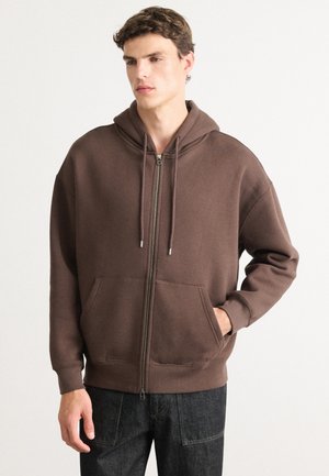 Brun zip-up hettegenser med kengurulomme, snorhette og ribbestrikkede ermer. Mykt stoff med glatt tekstur og minimal detaljer.