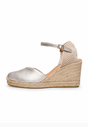 Sandalia cuña de esparto plateada con punta cerrada, parte trasera tejida, correa en el tobillo, hebilla dorada y plataforma con tacón de yute trenzado.