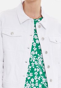 Chaqueta de denim blanca con botones plateados, con dos bolsillos en el pecho y un cuello clásico, superpuesta sobre un vestido floral verde.