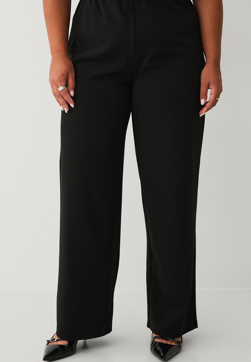 Pantalones negros de pierna ancha hechos de tela suave con cintura elástica, combinados con tacones negros puntiagudos. Sin patrones ni acentos visibles.
