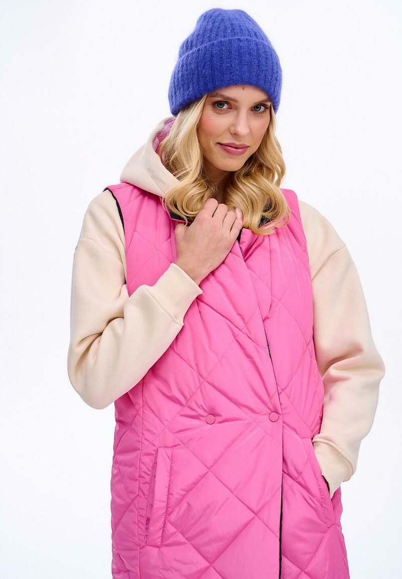 Růžová prošívaná vesta s béžovou kapucí, zdobená diagonálními vzory, bočními kapsami a černými akcenty. Modelka má na sobě modrou čepici.