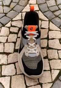 Musta ja beige tennari, jossa on tekstuurinen neulepäällinen, harmaat yksityiskohdat ja kontrastinen oranssi kantapään osa. Varustettu valkoisilla nauhoilla ja logoyksityiskohdalla.