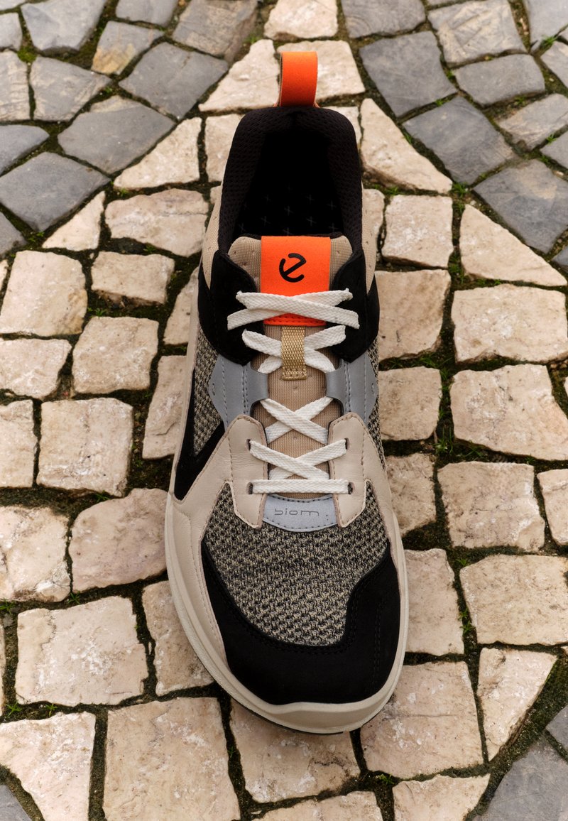 Schwarze und beige Sneaker mit einem strukturierten Strickobermaterial, grauen Akzenten und kontrastierendem orangefarbenem Fersenpatch. Mit weißen Schnürsenkeln und Logodetails. 