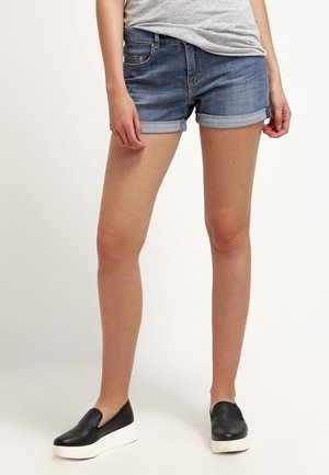 Person iført opkørte cowboyshorts, grå t-shirt og sorte slip-on sko med hvide såler, stående på en lys baggrund.