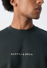 T-shirt en coton vert foncé avec un col rond classique, arborant un logo brodé argenté "SCOTCH & SODA" sur la poitrine.
