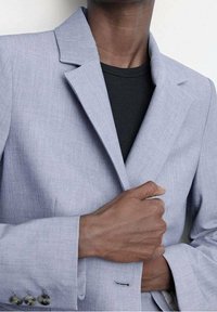 Light gray blazer with a textured fabric, notched lapels, single button closure, and silver buttons on the cuffs. Worn over a black shirt.