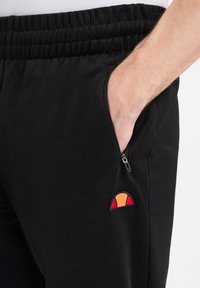 Schwarze Jogginghosen mit elastischem Bund, Reißverschluss-Tasche und einem rot, orange und gelb gestickten Logo an der Hüfte. Glatte Stoffstruktur.