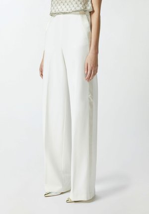 Pantaloni a gamba larga in tessuto bianco, caratterizzati da una texture liscia. Le cuciture laterali hanno una lucentezza, accentuata da dettagli metallici appuntiti sulle scarpe.