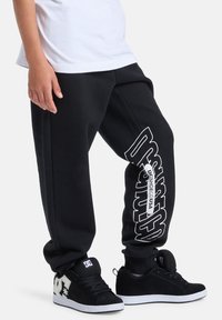 Schwarze Jogginghose mit lockerem Sitz, versehen mit weißem Markentext entlang des linken Beins. Kombiniert mit schwarzen Sneakern mit weißem Logo.