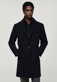 Black overcoat with a notch lapel, single button closure, and side pockets. Worn over a black zippered inner layer. Smooth texture.