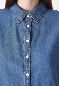 Gros plan d'une personne portant une chemise en denim bleu avec des boutons blancs et un col classique.
