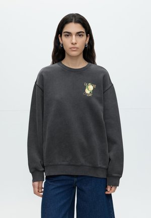 Kvinde med langt mørkt hår iført en sort oversized sweatshirt med gul pære-grafik og blå jeans, stående mod en hvid baggrund.