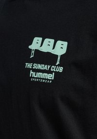 Svart T-shirt med en ljusgrön grafik av tre stiliserade stolar med texten "THE SUNDAY CLUB" och "hummel SPORTSWEAR" nedanför.