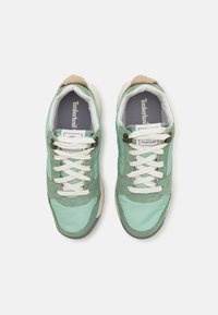Grüne und beige Sneakers aus einer Kombination von Wildleder und synthetischen Materialien, mit weißen Schnürsenkeln und einem grauen Innenetikett.