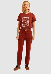 Camiseta de color óxido con un diseño gráfico blanco, combinada con pantalones de tiro alto a juego en color óxido y zapatillas de deporte con un patrón negro y marrón.