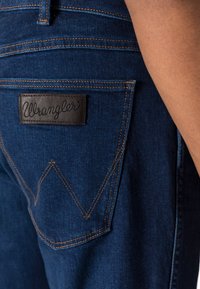 Vaqueros de denim azul oscuro con costuras naranjas en contraste, una etiqueta de cuero de Wrangler en el bolsillo trasero y un diseño distintivo en los bolsillos.