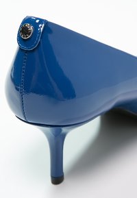 Chaussure à talon haut bleue brillante avec un talon étroit, des coutures visibles et un petit bouton métallique rond à l'arrière.