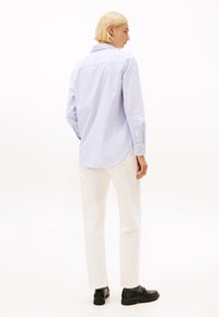 Person mit kurzen blonden Haaren, die ein hellblau gestreiftes Hemd, weiße Hose, graue Socken und schwarze Slipper trägt und mit dem Rücken zur Kamera steht.