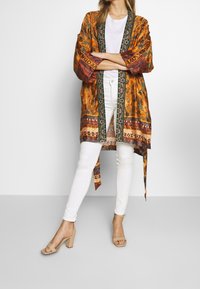 Kimono naranja con estampado y mangas anchas, combinado con una blusa blanca y jeans blancos, acompañado de sandalias de tacón bloque nude.