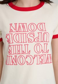 T-shirt en coton crème avec col côtelé rouge. Présente un texte rouge indiquant "WELCOME TO THE UPSIDE DOWN" dans un design audacieux et inversé.