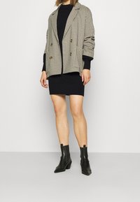 Blazer croisé à motif à carreaux beige et noir, associé à une robe noire ajustée et des bottines noires à talon large.