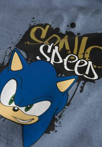 Plava pamučna majica s karikaturom koja ima zelene oči, žute akcentne boje i natpis "Sonic Speed" u crnom i zlatnom grafitnom stilu.