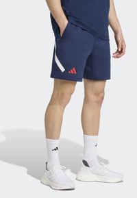 Granatowe szorty sportowe z białym paskiem i czerwonym logo; noszone z białymi skarpetkami i trampkami. Miękka tkanina, luźny krój.