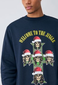 Marinblå sweatshirt med grafik föreställande dödskallar i tomteluva och texten "WELCOME TO THE JINGLE" ovanför en korsdesign.
